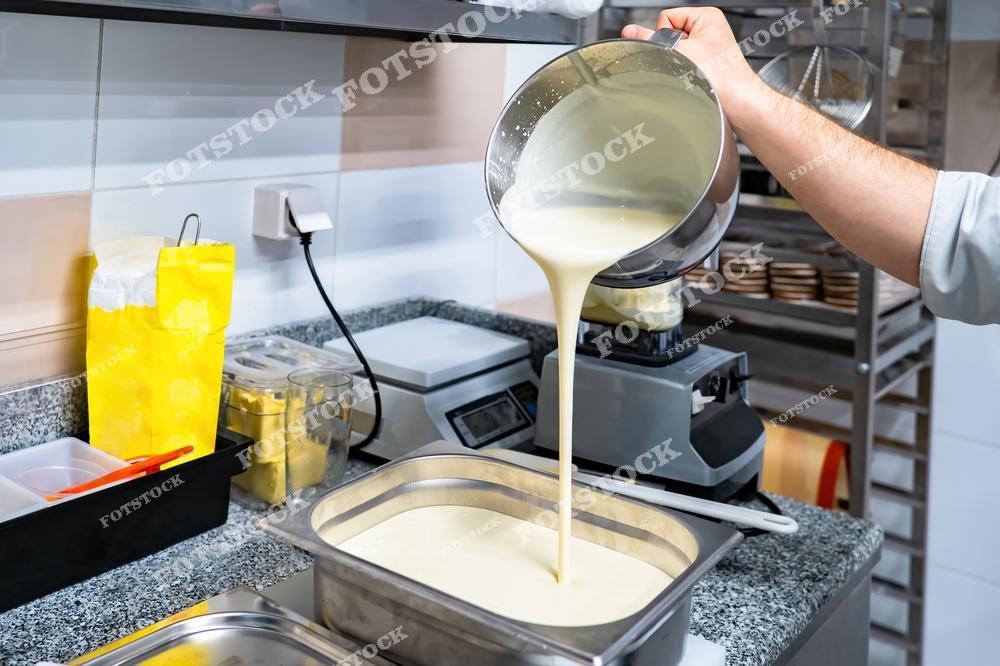 На фотографии изображен процесс приготовления на профессиональной кухне. Повар переливает жидкое тесто из миски в контейнер на рабочей поверхности. Снимок передает атмосферу ресторанной кухни и подчеркивает профессионализм в кулинарном искусстве.